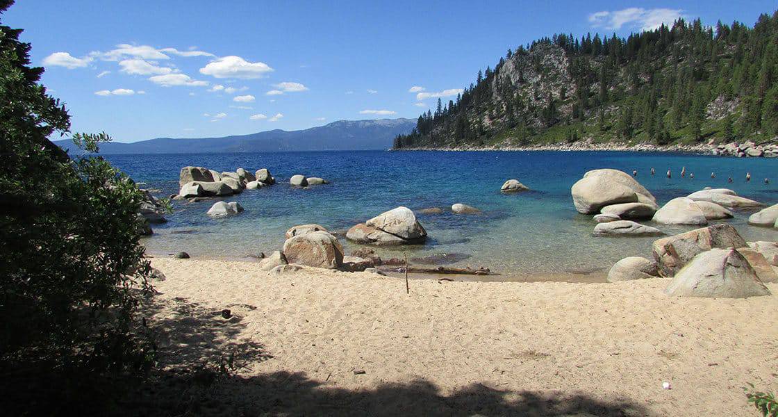Sandy shoreline at Skunk Harbor on Lake Tahoe's east shore with clear turquoise water, granite boulders, and pine forest climbing the cliffs behind