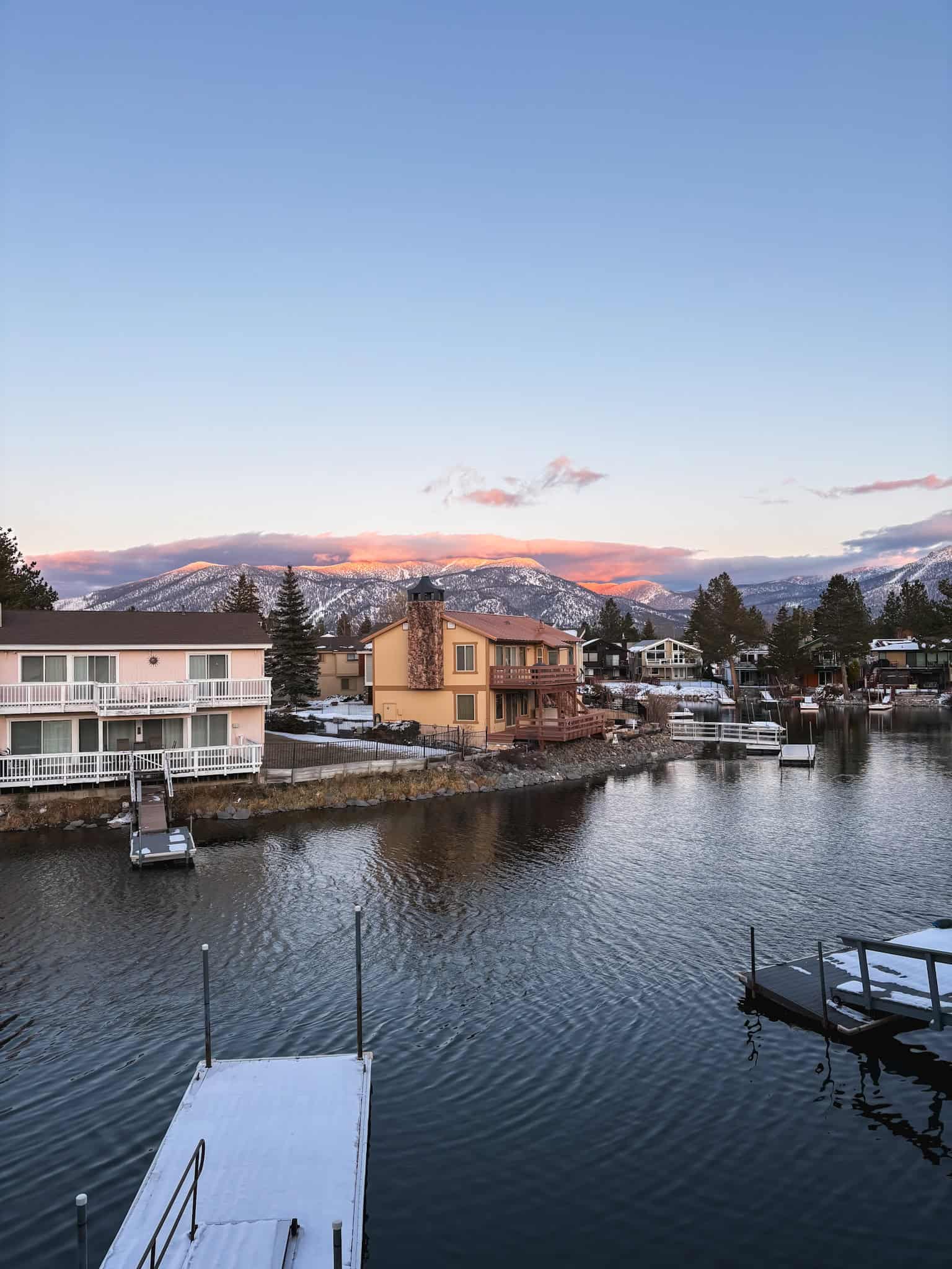 Winter sunrise turning the Sierra Nevada peaks pink above a South Lake Tahoe neighborhood canal with snow-dusted docks and calm water