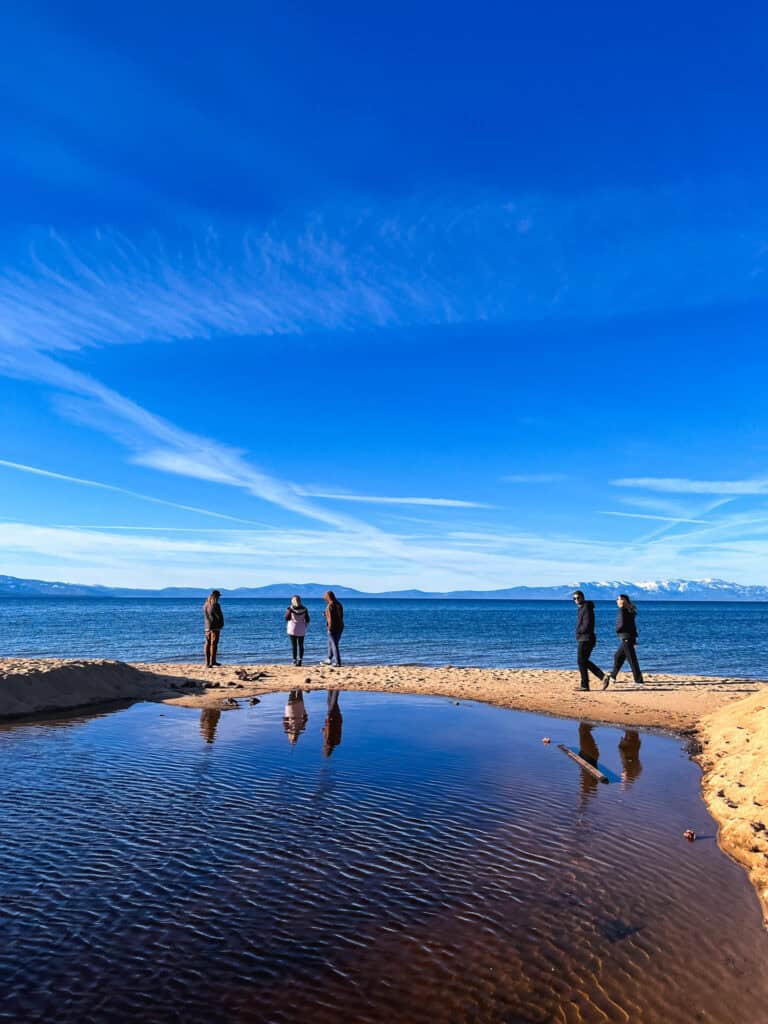 Group of people walking along a sandy Lake Tahoe beach in winter with their reflections in a shallow tide pool and snow-capped mountains across the water