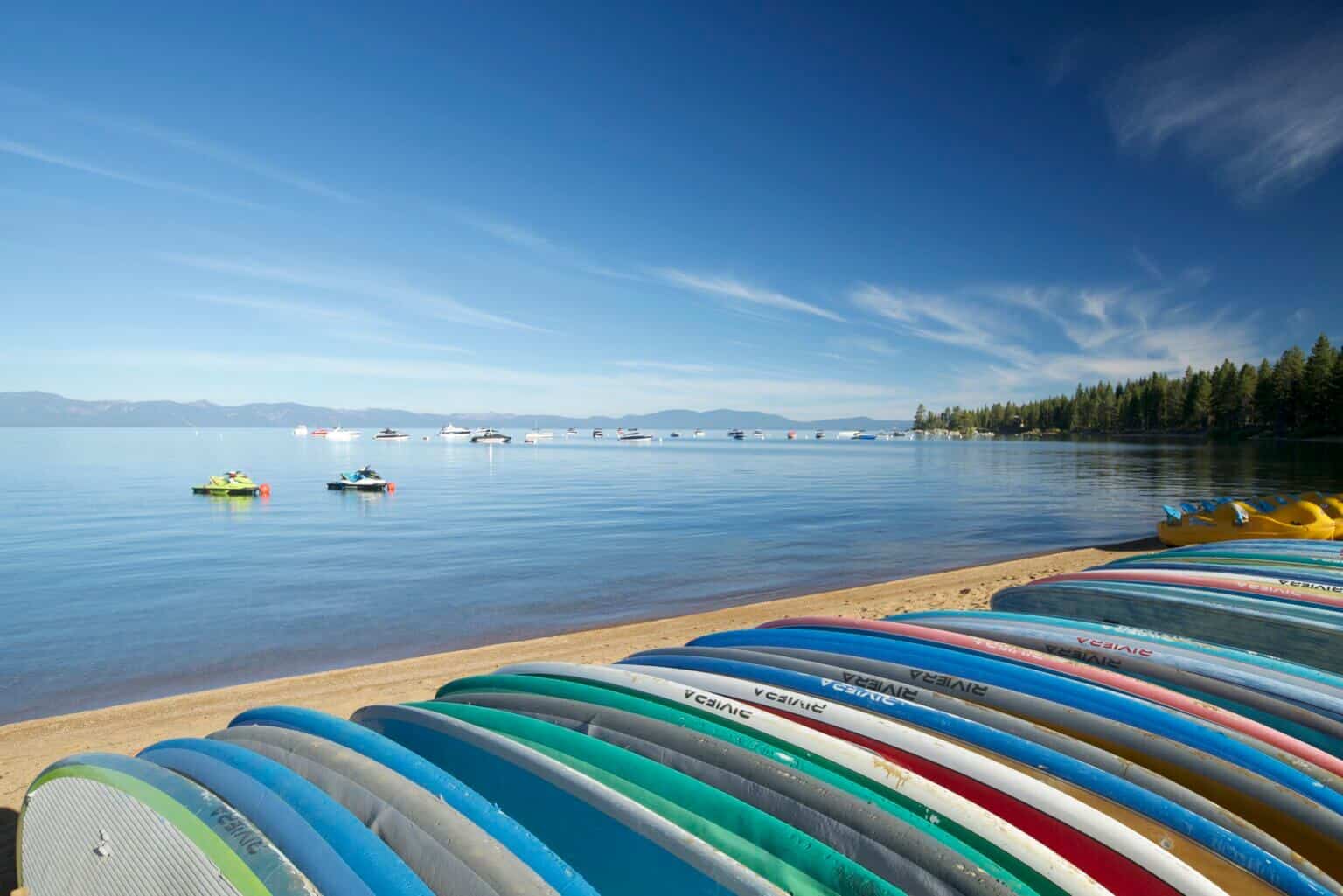 Rows of colorful paddleboards lined up on the sand at Zephyr Cove with jet skis and boats anchored on the calm lake and pine-forested mountains in the distance