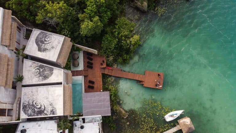 Drone view of Amainah hotel in Bacalar showing nautilus shell murals painted on rooftops, a narrow infinity pool, wooden dock extending over the turquoise lagoon, and a sailboat moored below