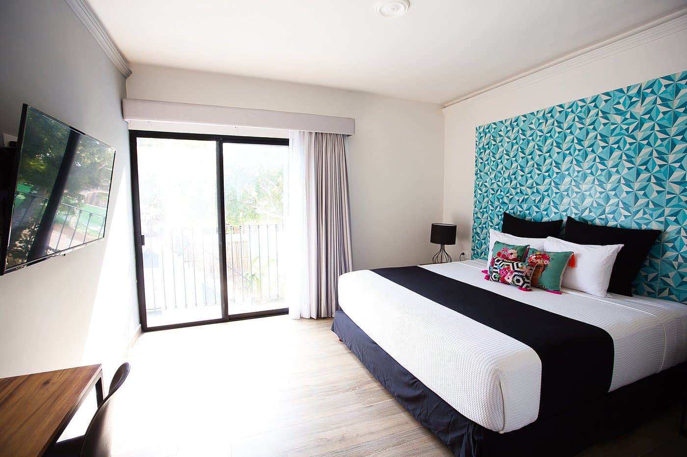 Bright hotel room at Chukum Bacalar with a teal geometric tile headboard, white bedding with colorful embroidered pillows, and a sliding glass door opening to a balcony