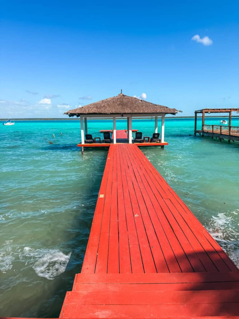Bright red dock extending over turquoise water at one of the best hotels in Bacalar, with a palapa-covered lounge platform and lounge chairs over the lagoon