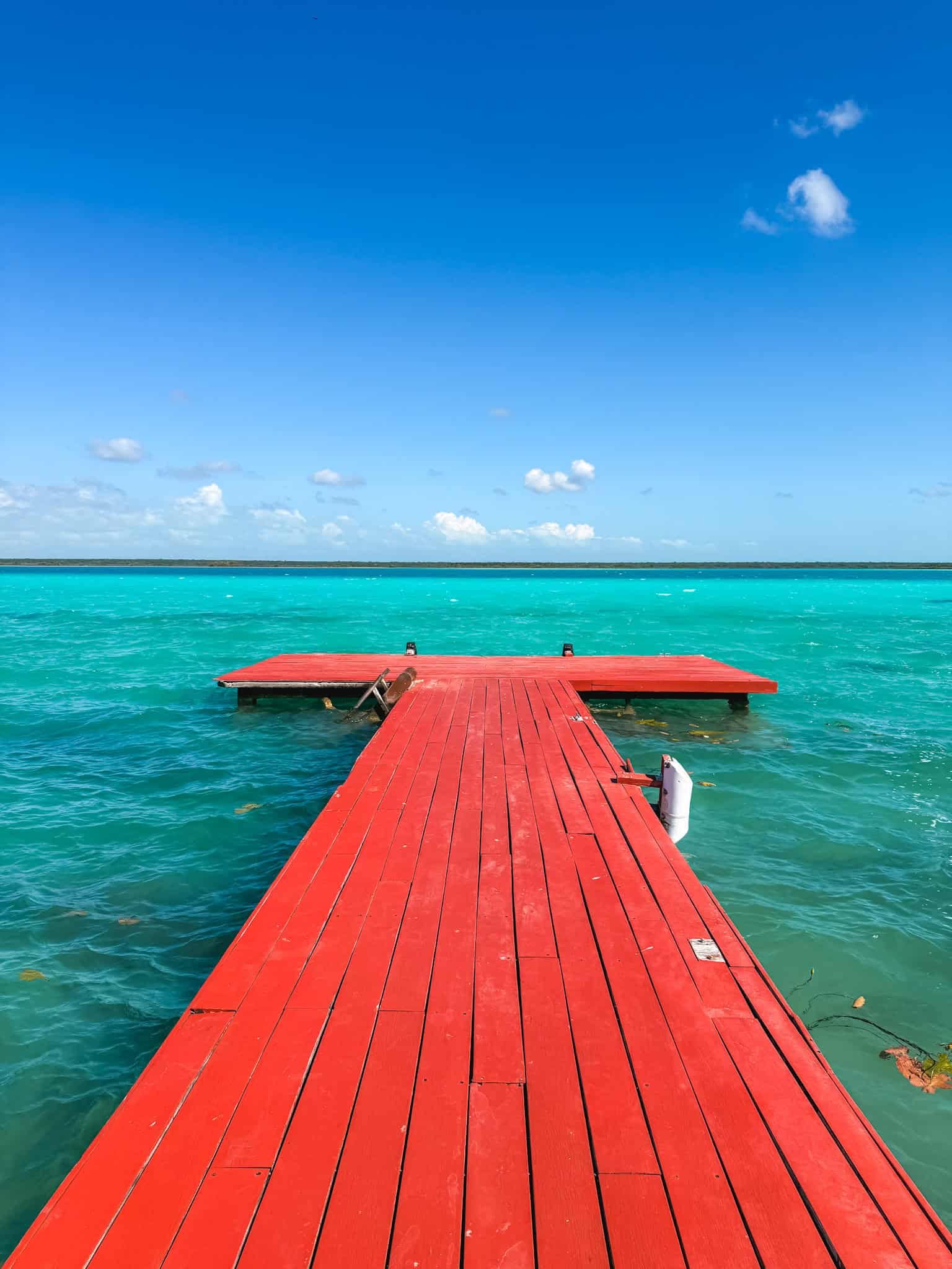 Red wooden dock stretching into the electric turquoise water of Bacalar lagoon on a clear sunny day, with a small swim platform at the end