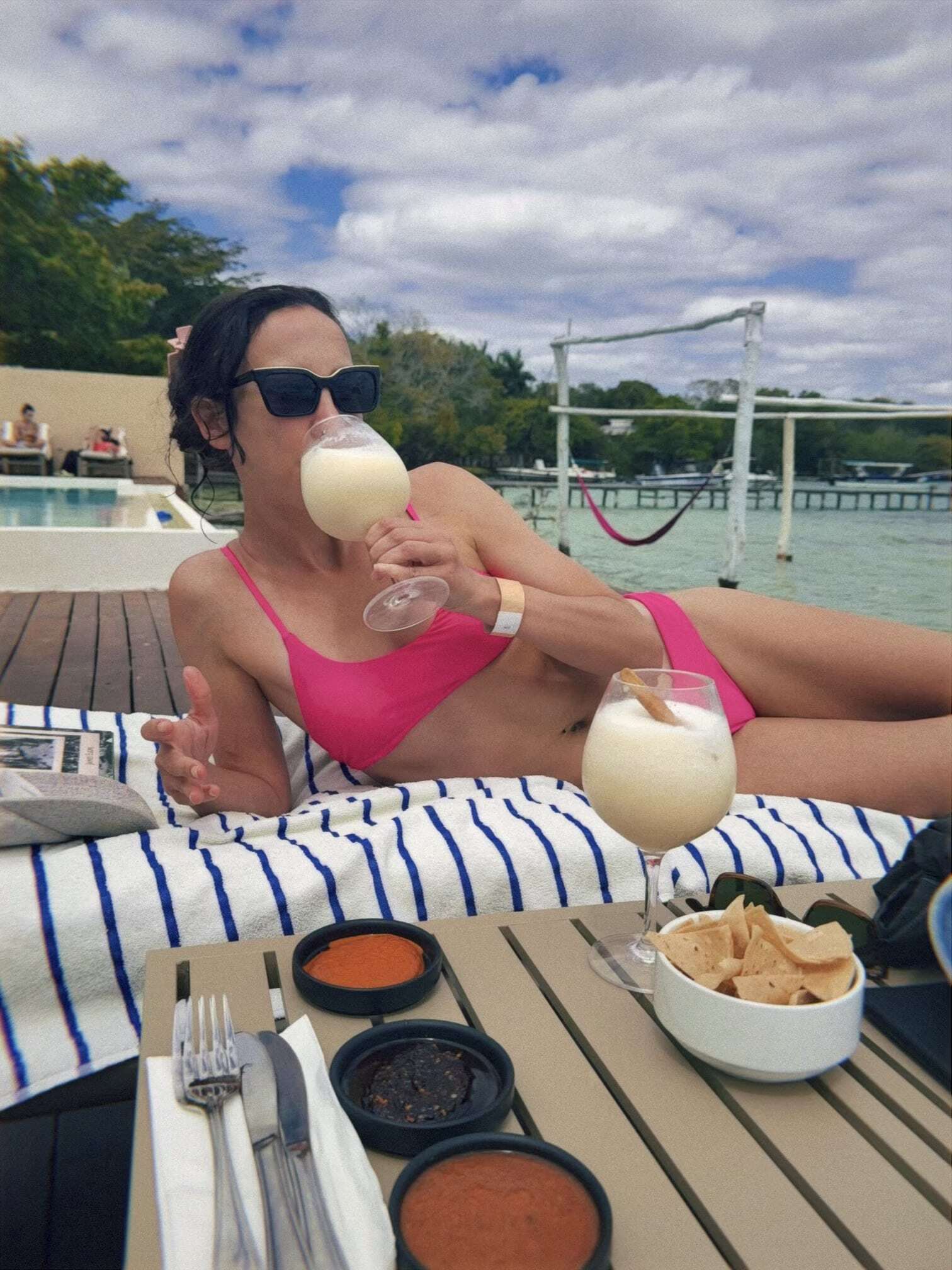 Woman in a pink bikini sipping a frozen margarita on a sun lounger at one of the best hotels in Bacalar, with chips and salsa on the side table and the turquoise lagoon in the background