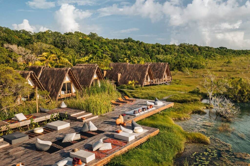 Aerial view of Our Habitas Bacalar, one of the best hotels in Bacalar, with A-frame thatched bungalows nestled in jungle greenery alongside a wooden deck lounge area next to the lagoon