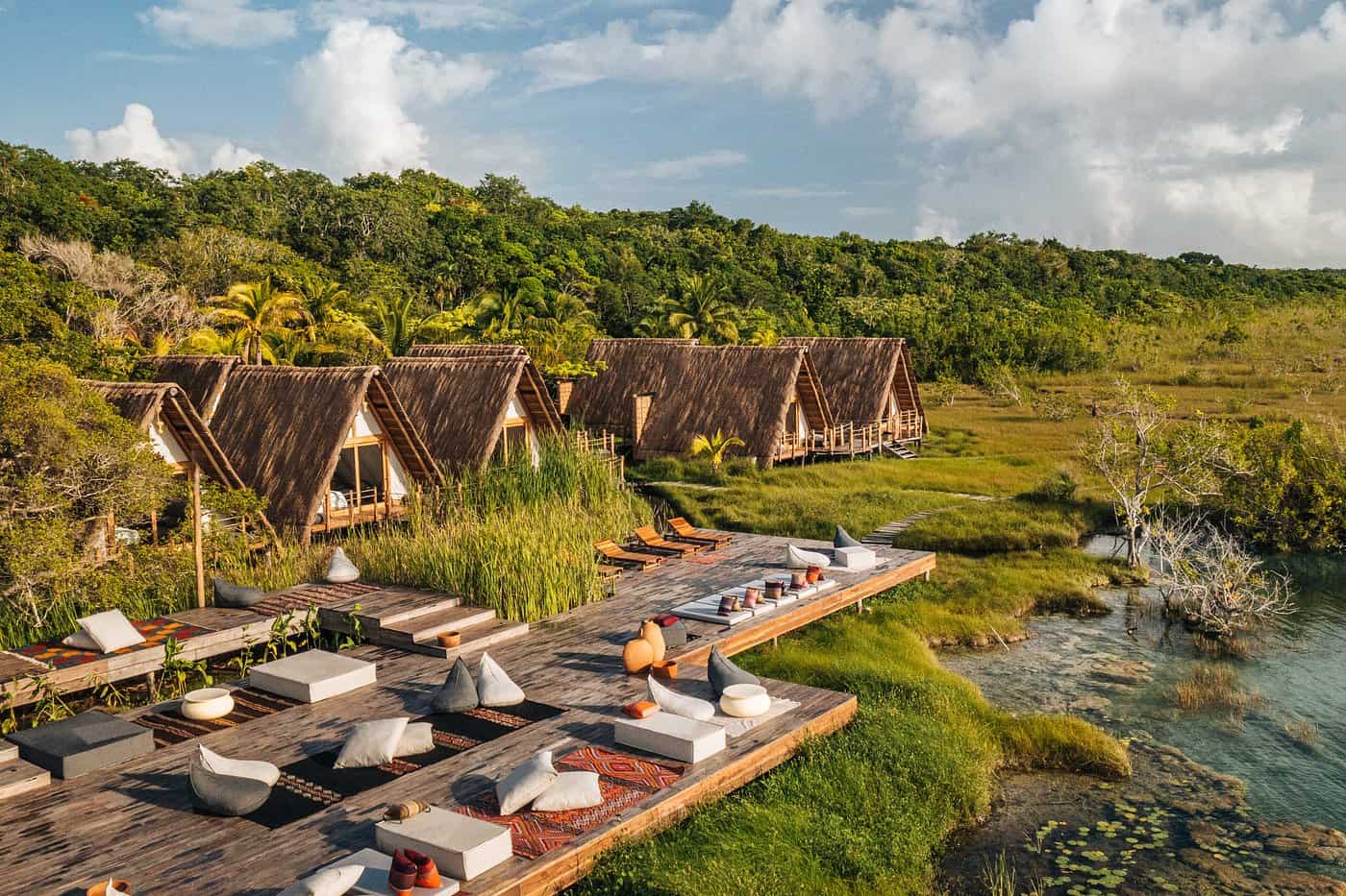 Aerial view of Our Habitas Bacalar, one of the best hotels in Bacalar, with A-frame thatched bungalows nestled in jungle greenery alongside a wooden deck lounge area next to the lagoon