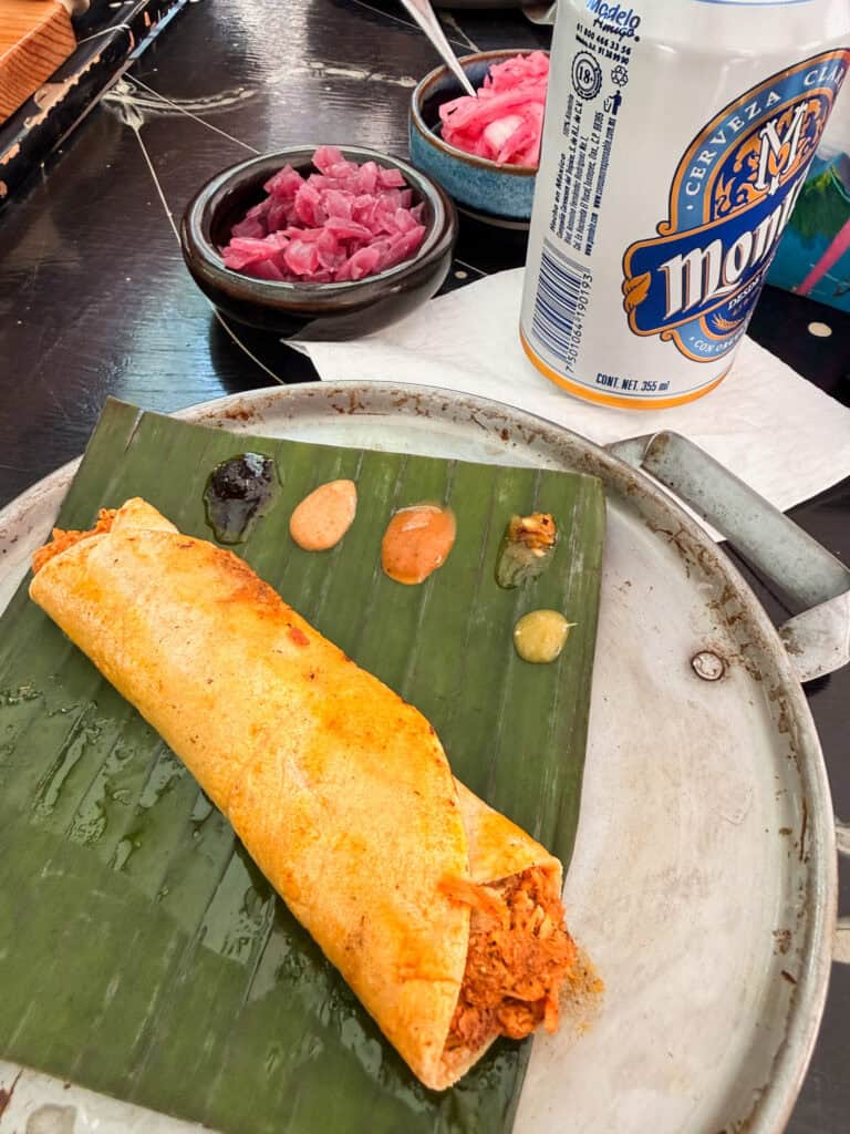 A crispy rolled taco served on a banana leaf with four dipping sauces and pickled red onion on a Mexico City food tour, with a Modelo can in the background