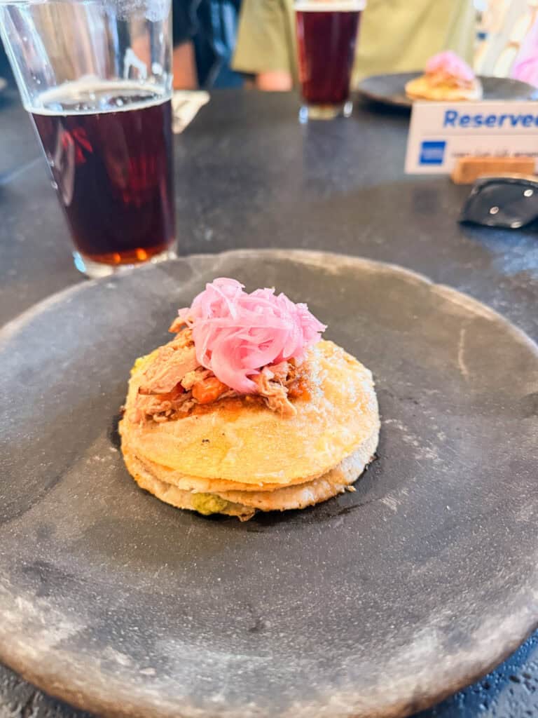 A pork taco topped with pickled pink onions on a dark stone plate during a Mexico City food tour, served with agua de jamaica in the background