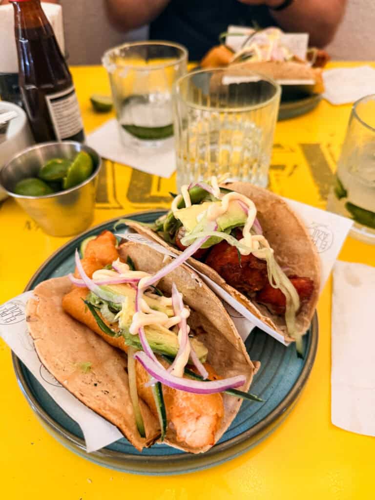 Two shrimp tacos with avocado, pickled red onion and chipotle mayo on whole wheat tortillas served on a teal plate during a Mexico City food tour, with fresh limes on the side