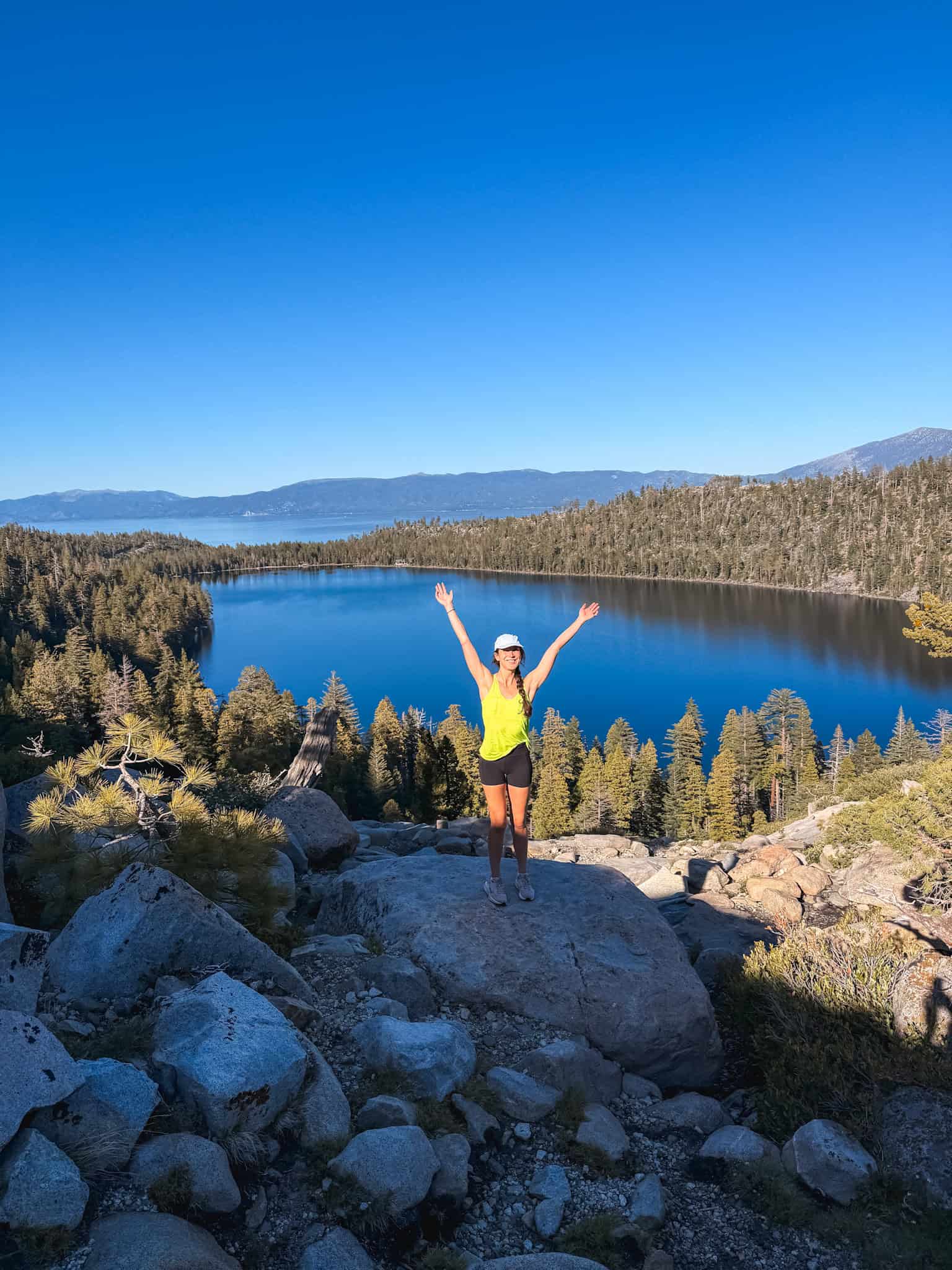 Woman with arms raised standing on a granite boulder above Cascade Lake with Lake Tahoe visible in the background on a clear blue sky day