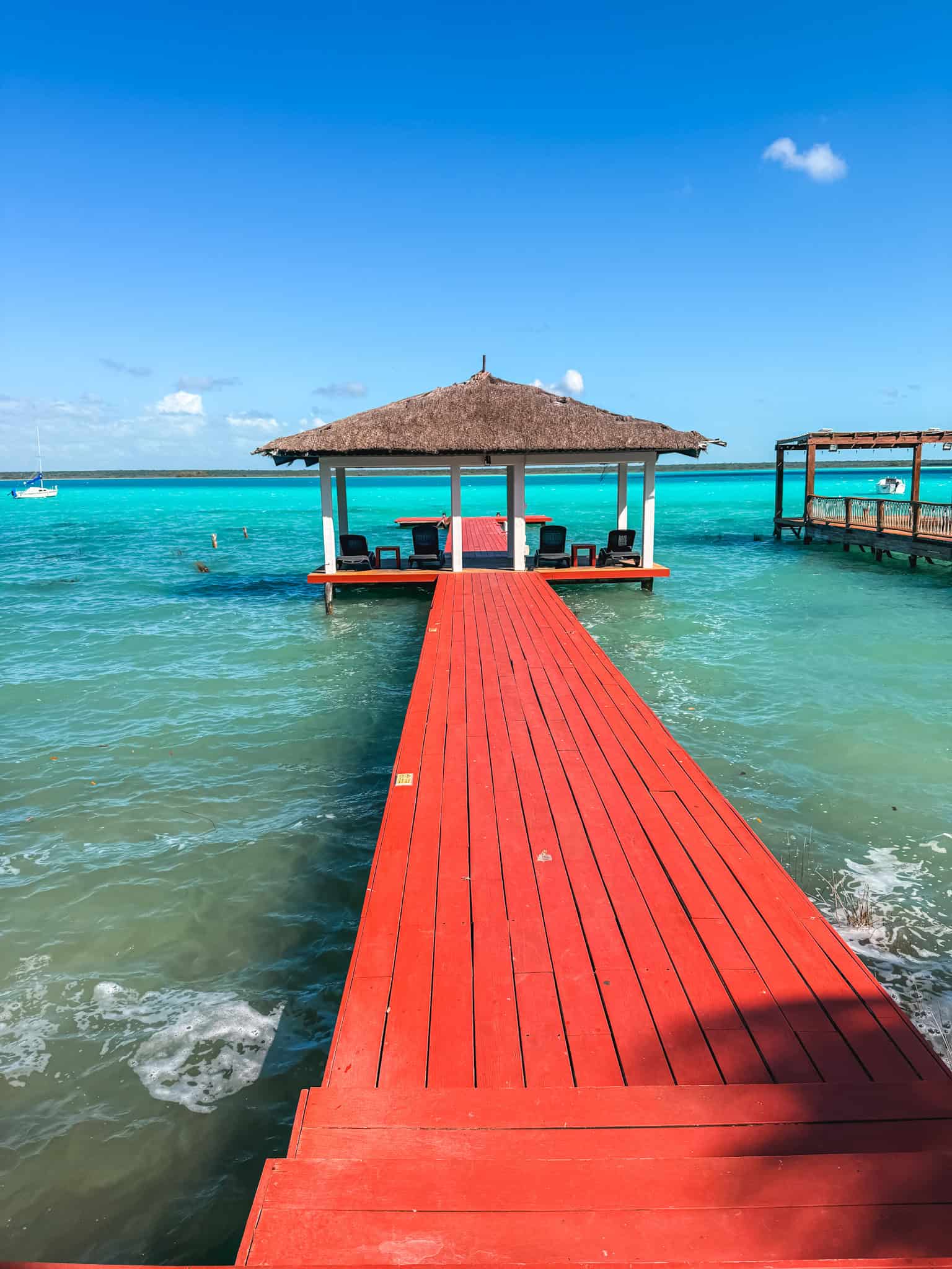 Bright red wooden pier extending over the turquoise waters of Laguna Bacalar at a beach club, with a thatched gazebo at the end and a sailboat in the distance