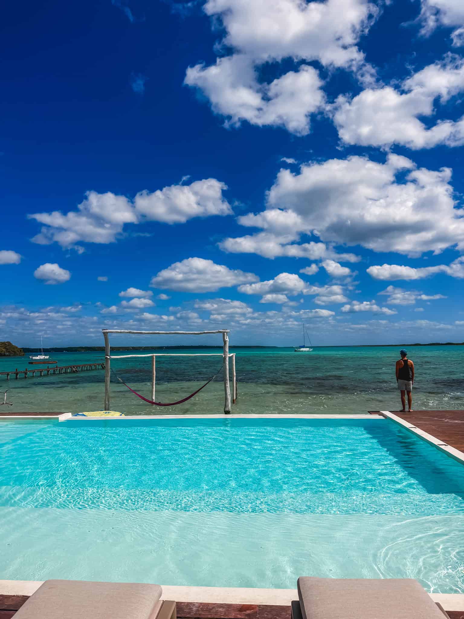 Infinity pool at a Bacalar beach club overlooking the lagoon, with a hammock frame and sailboat visible on the turquoise water