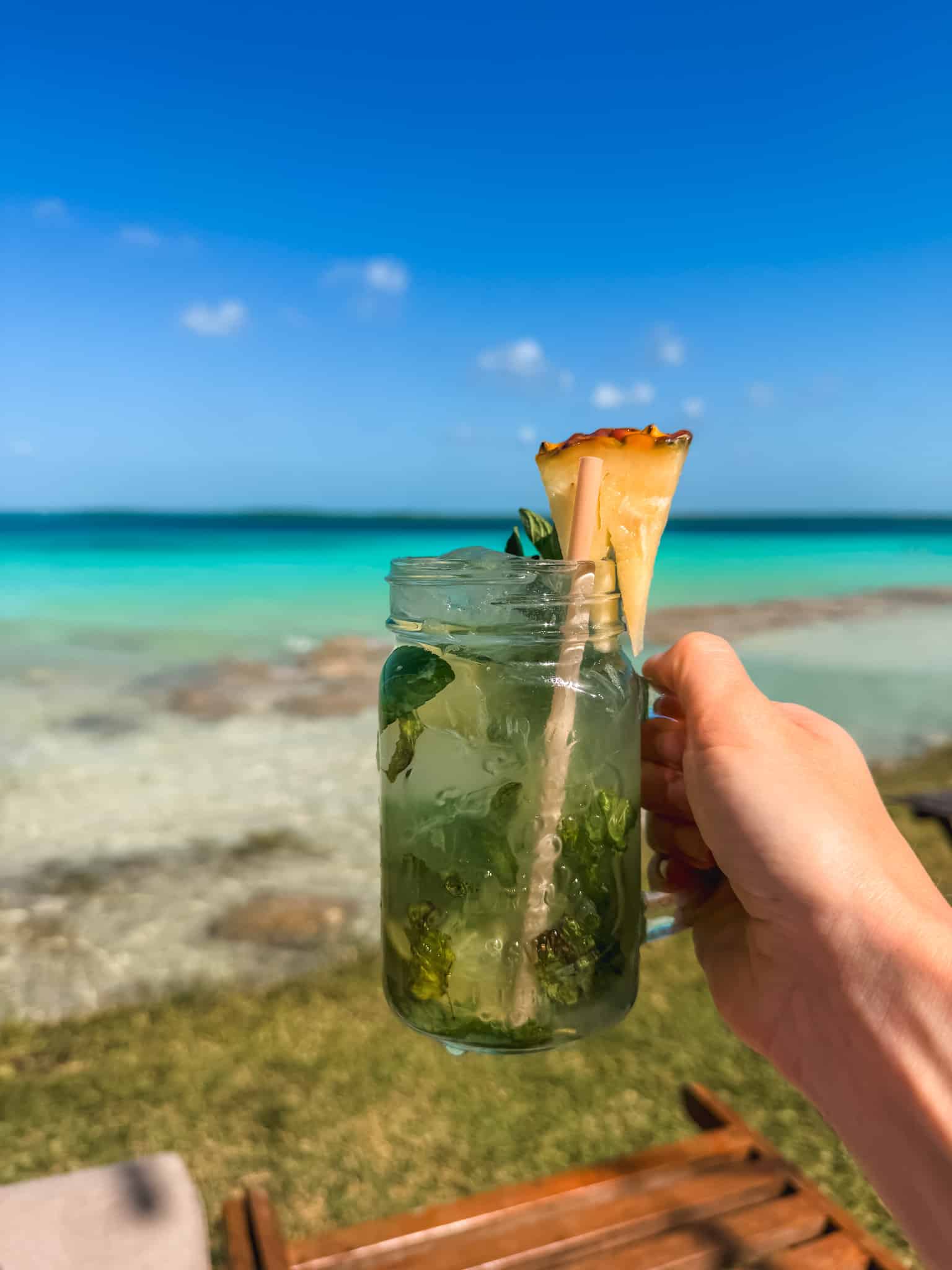 Hand holding a mojito in a mason jar with a pineapple garnish at a Bacalar beach club, with the turquoise lagoon glowing behind it