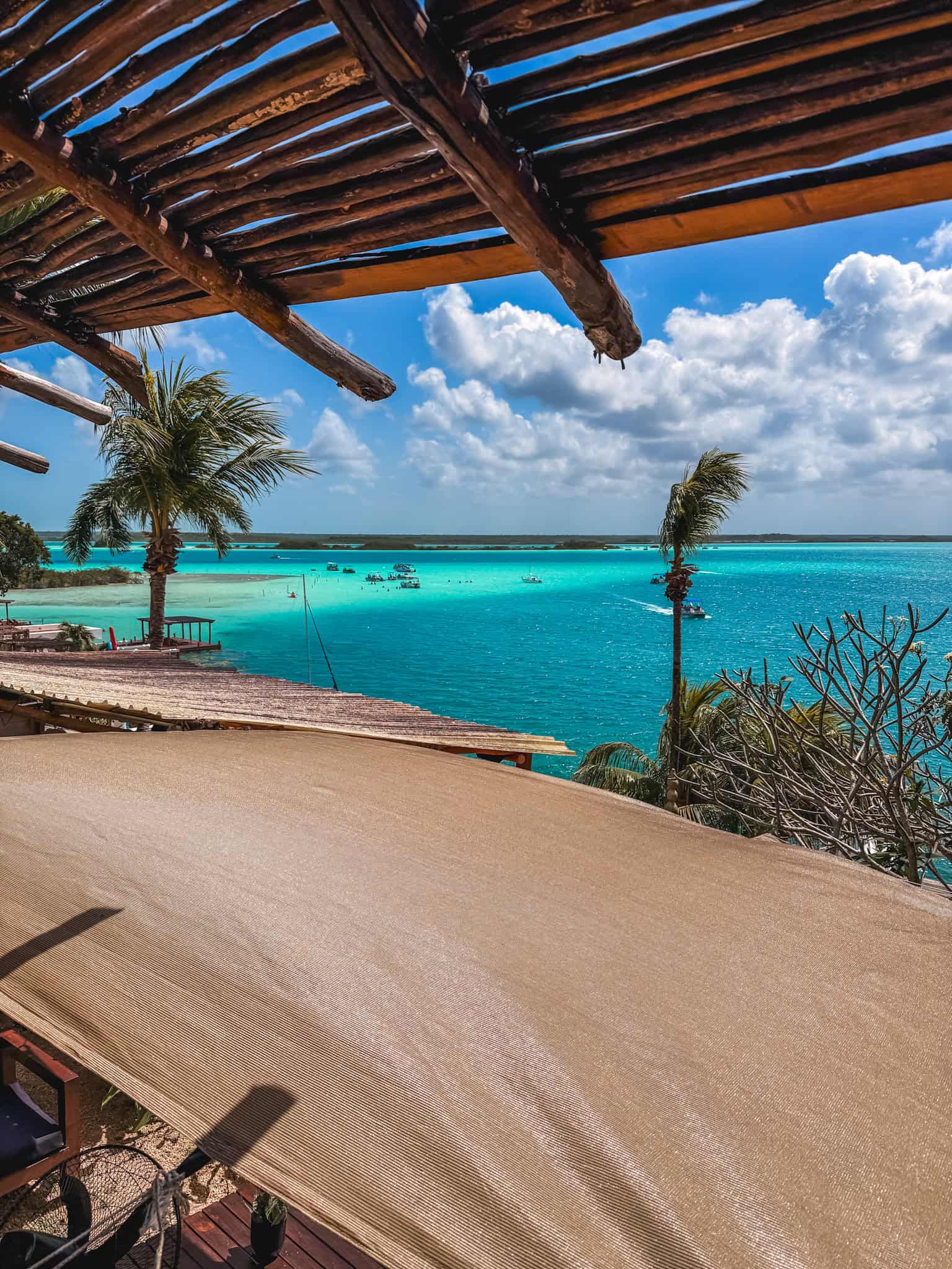 View of Bacalar Lagoon's famous multi-colored turquoise waters from a rooftop palapa bar, with sailboats dotting the horizon