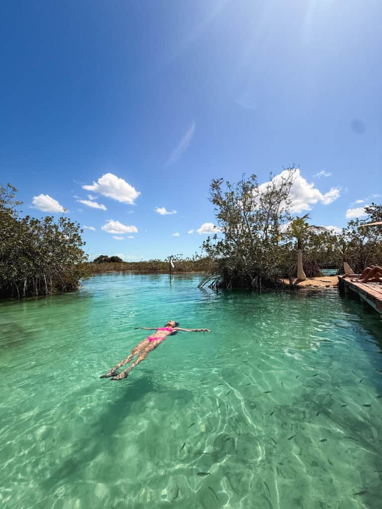 Woman floating face-up in the clear turquoise water of Los Rapidos in Bacalar, surrounded by mangroves on a sunny day