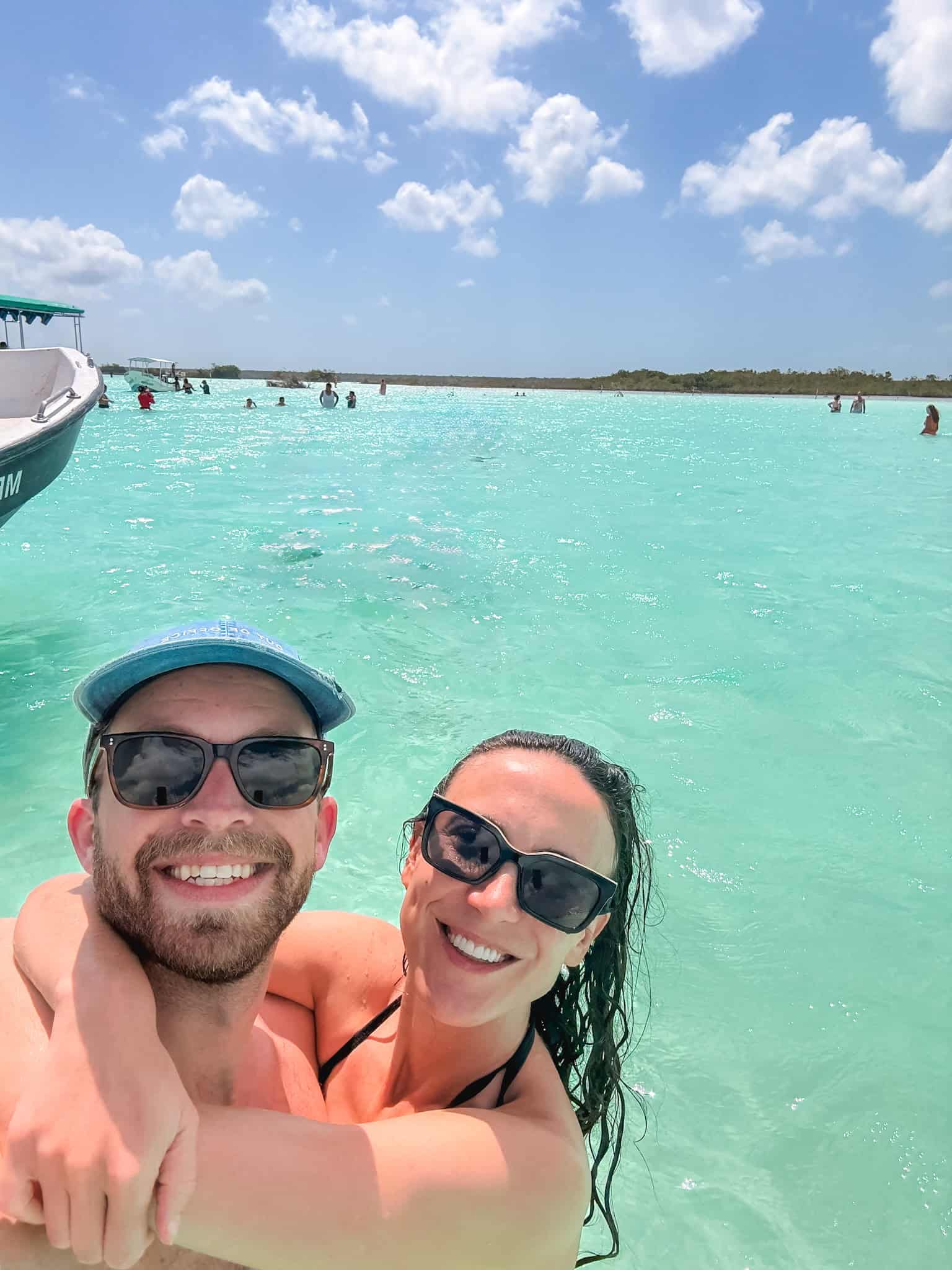 Couple smiling for a selfie while swimming in the turquoise waters of Laguna Bacalar during a boat tour, with other swimmers in the background