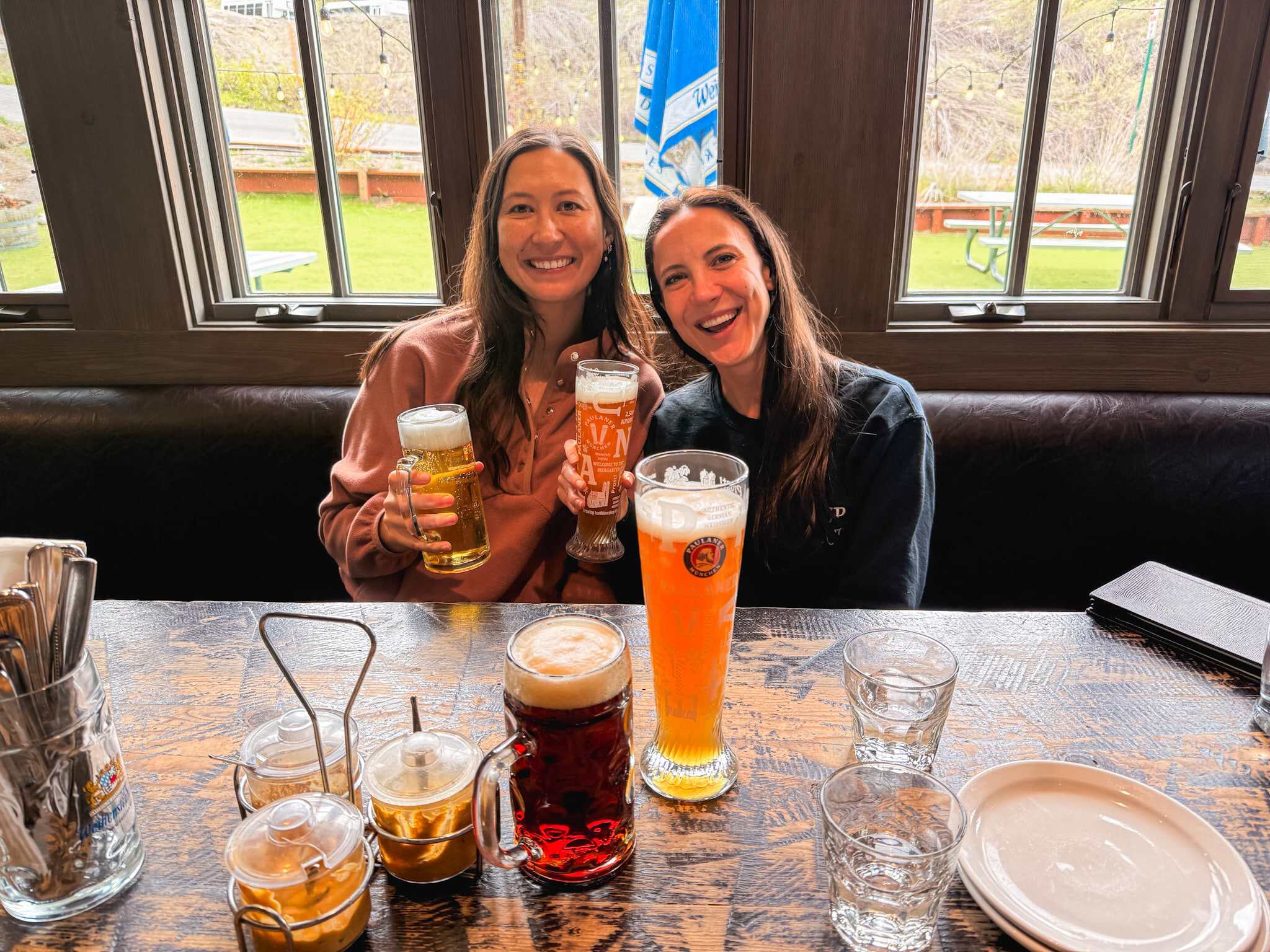 Two women smiling and holding German beer steins at a Lake Tahoe brewery, with a Paulaner wheat beer and dark beer on the table