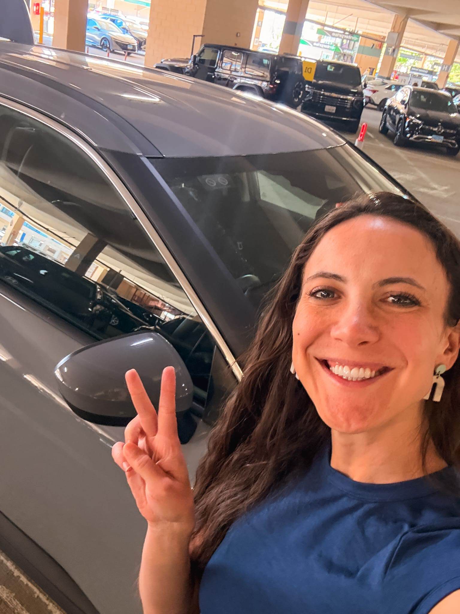 Woman giving a peace sign next to a rental car in a parking garage, picking up her rental car for a Lake Tahoe road trip