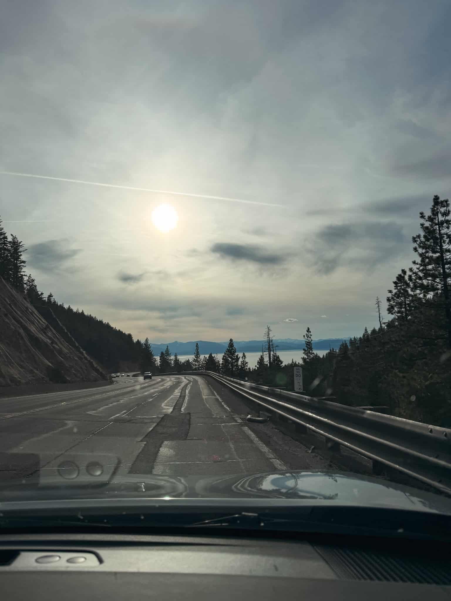 View through a car windshield of the highway approaching South Lake Tahoe, with pine trees and the lake visible in the distance on a cloudy day
