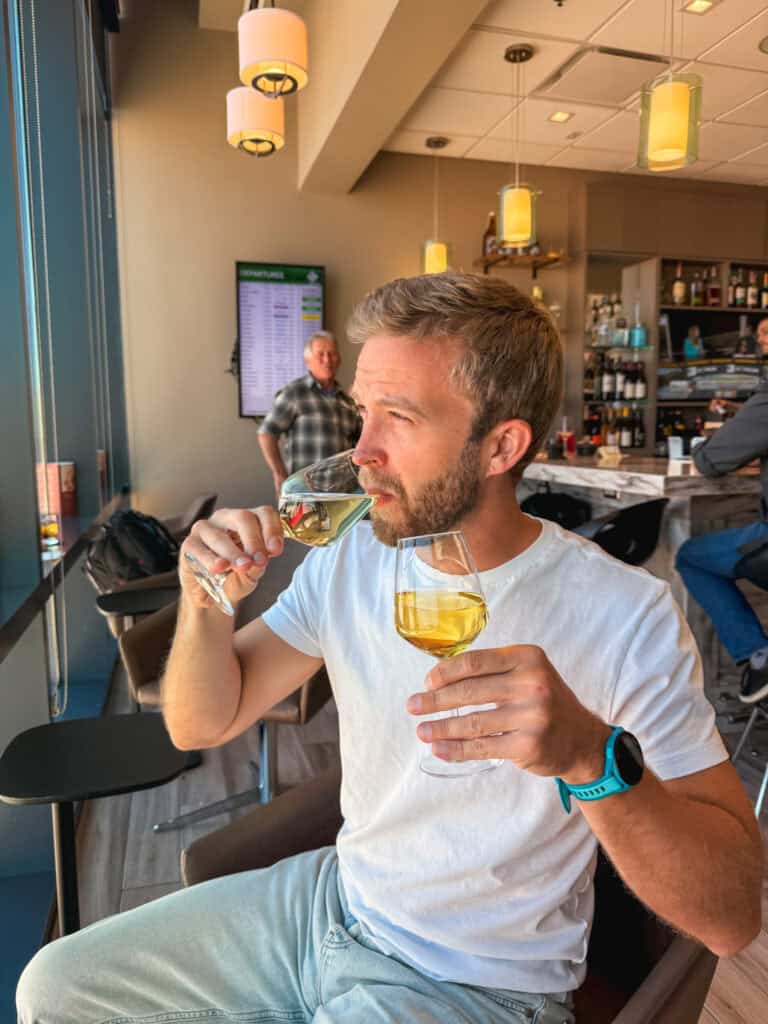 Man sipping white wine at the Escape Lounge inside Reno-Tahoe International Airport, with the full bar and departures board visible in the background