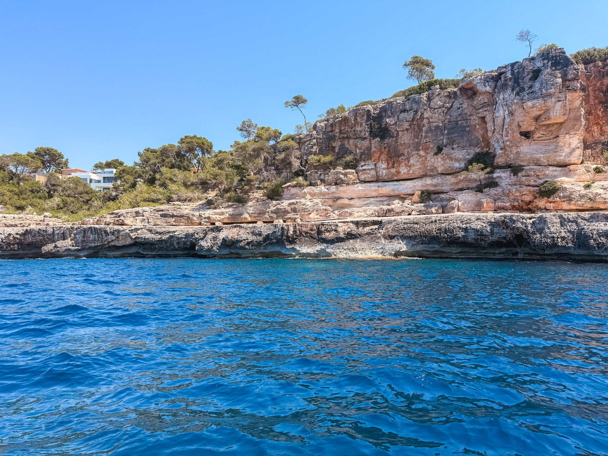 Turquoise water against limestone cliffs on a Mallorca boat tour, with a hillside villa peeking through the pine trees above.