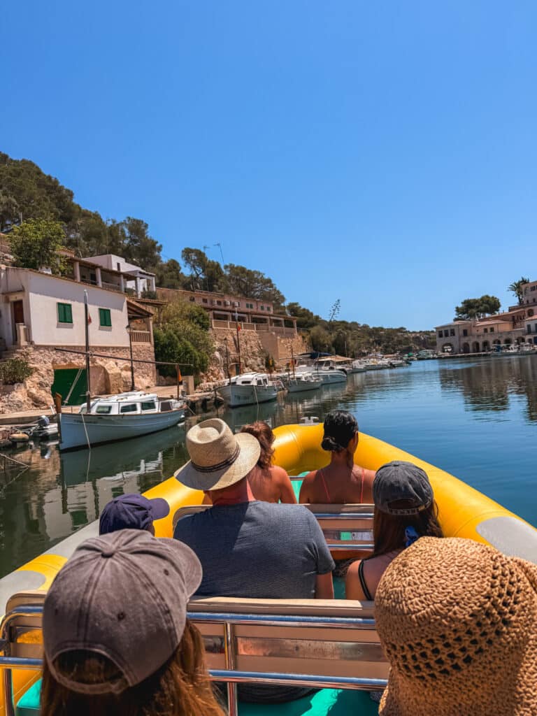 Passengers in sunhats riding through the canal at the start of a Mallorca boat tour, passing traditional fishing boats.
