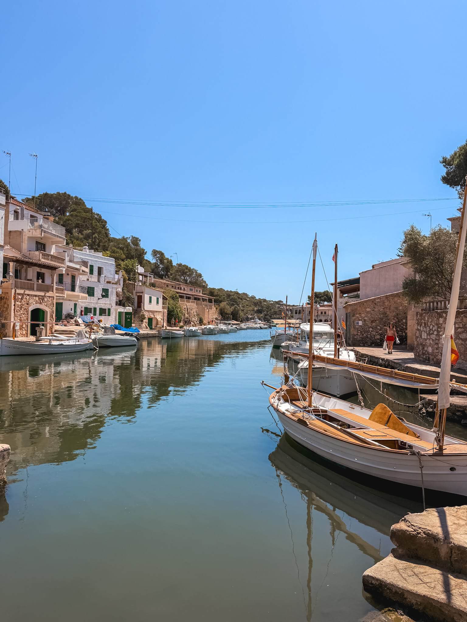 Traditional fishing boats moored in a quiet Mallorca harbor canal, lined with white stone houses.