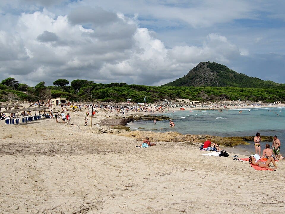 Wide sandy shoreline at Cala Agulla with sunbathers spread across the beach, pine forest and a rounded hill in the background