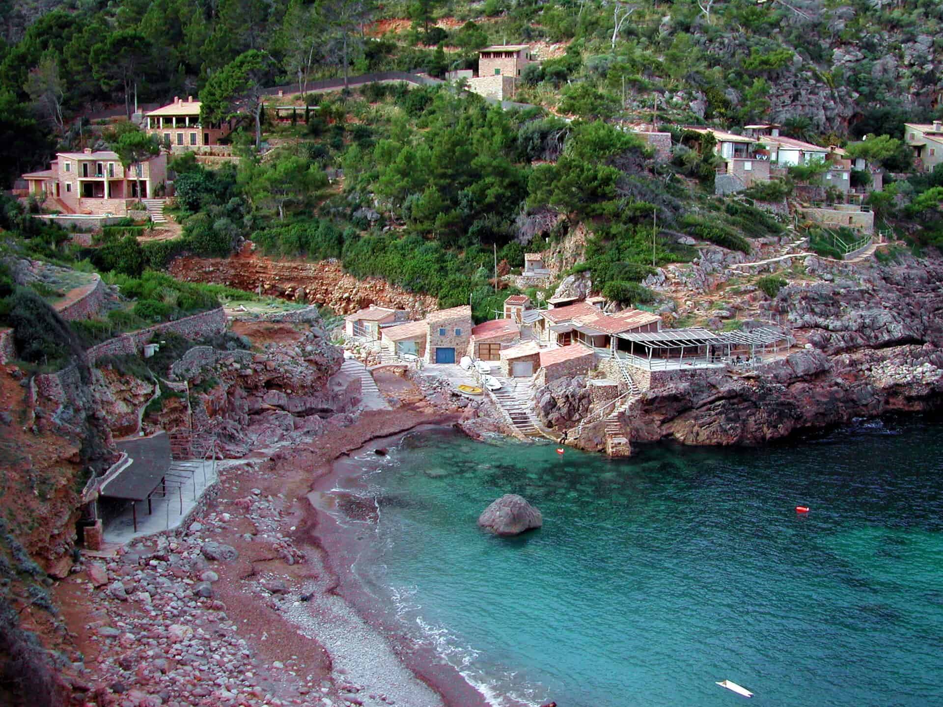 Small pebble cove below the cliffside village of Deià, stone fishing huts lining the red tinted rocks
