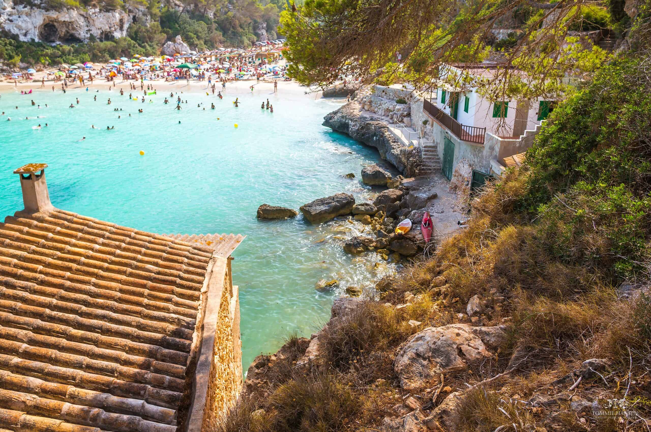 Crowded turquoise cove at Cala Llombards, swimmers filling the water beneath whitewashed stone buildings