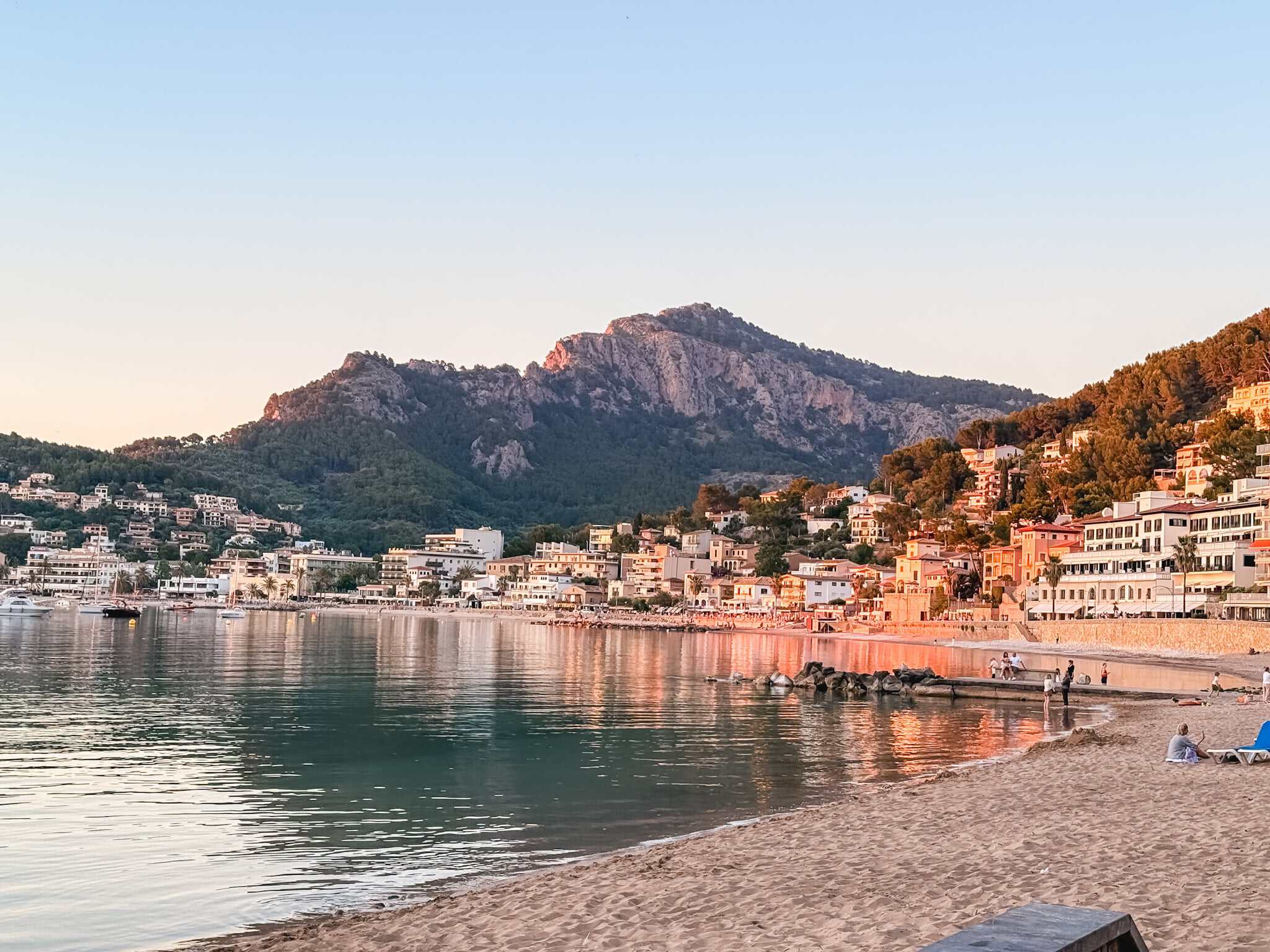 Port de Soller beach at golden hour, mountains rising behind a curved bay lined with waterfront buildings